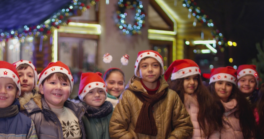 4k Happy children are jumping , laughing  and looking camera into front of the beautiful house decorated with lights and garlands.  Portrait of many kids with Santa hat and costumes partying at  night Royalty-Free Stock Footage #1095827543