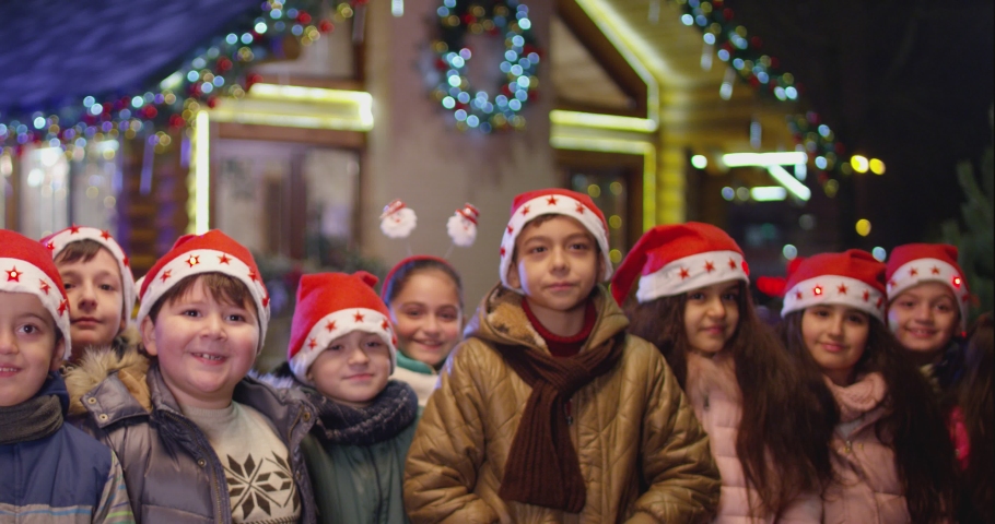 4k Happy children are jumping , laughing  and looking camera into front of the beautiful house decorated with lights and garlands.  Portrait of many kids with Santa hat and costumes partying at  night Royalty-Free Stock Footage #1095827557