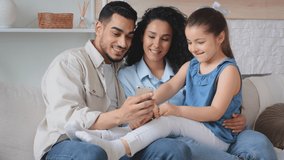 Multiracial multiethnic happy family Hispanic Arabian Caucasian parents with small daughter child girl sitting on couch watching funny video film movie cartoons in mobile phone video call fun laugh