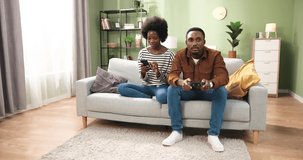 African American young handsome man sitting on sofa at home playing video games on console having fun while his wife typing and browsing online on mobile phone and showing him something Family concept