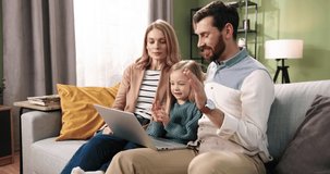 Joyful Caucasian young family parents and small adorable child videochatting talking on video call online on laptop waving their hands and smiling. Family time together. Home concept