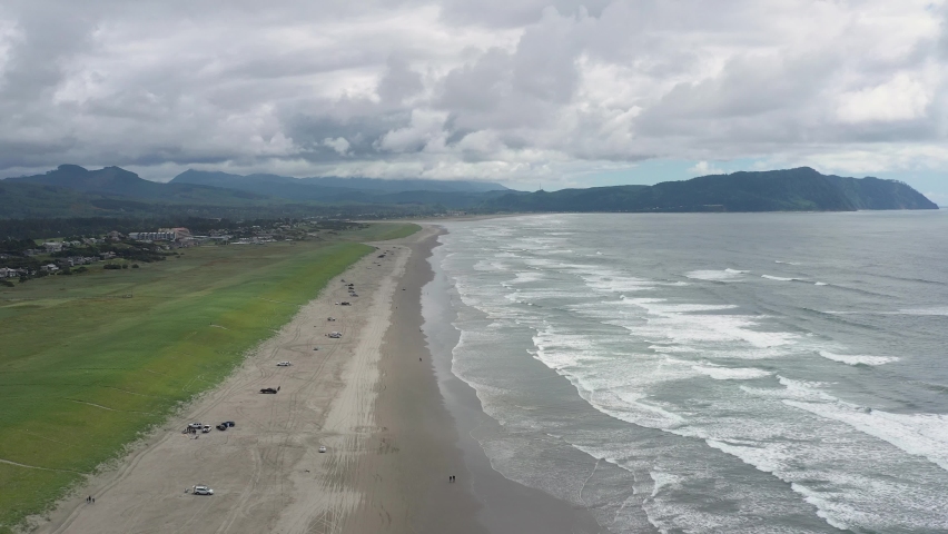 Aerial drone flight high above people with cars having fun activities at the beach in Oregon as ocean waves wash on the shore.