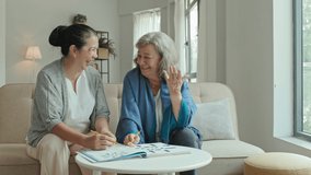 Medium tracking shot of cheerful Asian and Caucasian elderly women sitting on sofa in nursing home, chatting and laughing while solving crossword together - Powered by Shutterstock - Get 15% off with code: PIKWIZARD15
