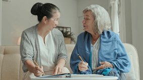 Medium shot of two positive Asian and Caucasian elderly women sitting on sofa in nursing home, chatting and smiling while solving crossword puzzle together - Powered by Shutterstock - Get 15% off with code: PIKWIZARD15
