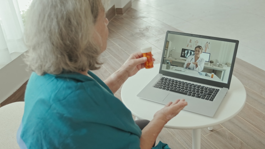 Over the shoulder shot of elderly woman sitting at home, holding pills and talking with female doctor via online video call on laptop | Shutterstock HD Video #1095845309