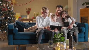A father sits with his children in the living room on the couch while they watching something on a tablet. The room is decorated in a festive spirit, as well as the coming Christmas.