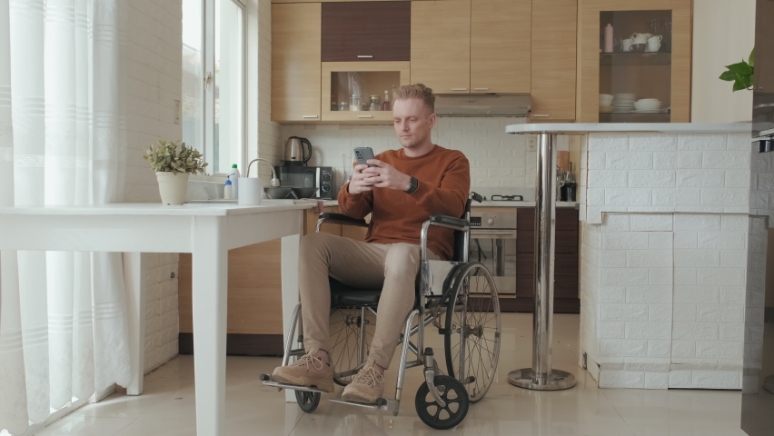Stab shot of young Caucasian man in wheelchair using his phone then smiling at camera at bright modern kitchen