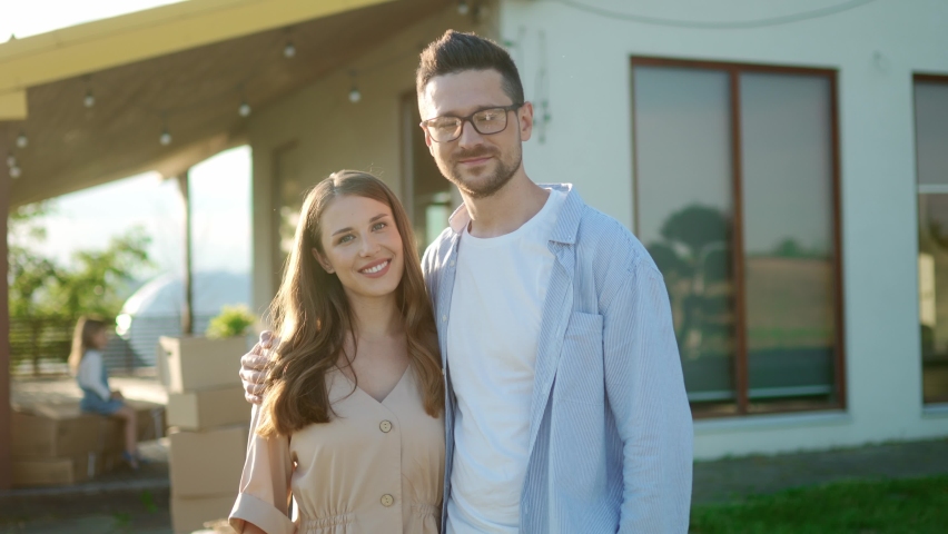 Close up portrait of happy young married couple smiling at camera while standing outside their new home. Kids with boxes on background. Cheerful Caucasian family outdoors. Family weekend concept