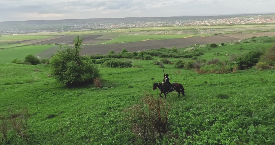 FPV sports drone shot traditional culture Caucasian horseman with gun riding horse at summer mountain valley landscape. Aerial view male equestrian highlander at picturesque dramatic sky rocky scenery