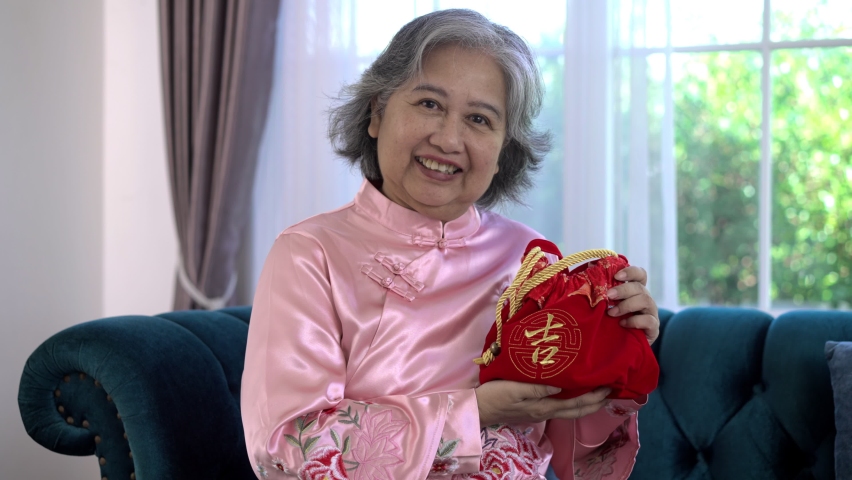 Handheld medium shot, Portrait Asian Senior woman wearing Chinese traditional costume showing lucky red bag in hand, She smiling and looking to camera, Chinese New Year concept