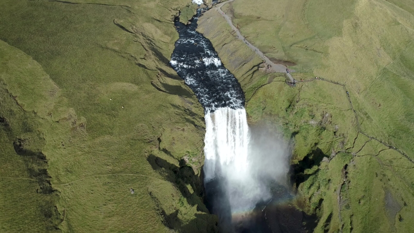 Aerial drone footage captured in Iceland near Skogafoss waterfall. Powerful river water falls down the cliff and cloud of water drops is covering the waterfall. In the cloud you can see small rainbow.