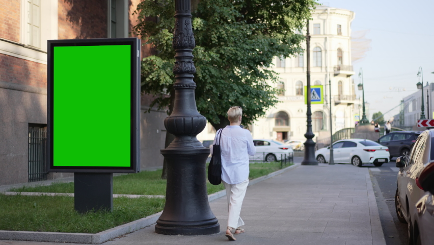 4k Green mockup billboard stands and woman walks in city spbd. Empty vertical display for advertising standing, cars driving along road and female walking outdoors on summer day.  