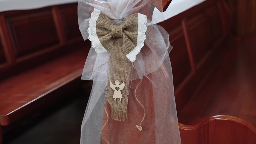Linen bow decorated with cotton lace and wooden angel is attached to the edge of empty wooden bench in the church, close-up. Festive decoration of the temple for Easter. | Shutterstock HD Video #1095872993