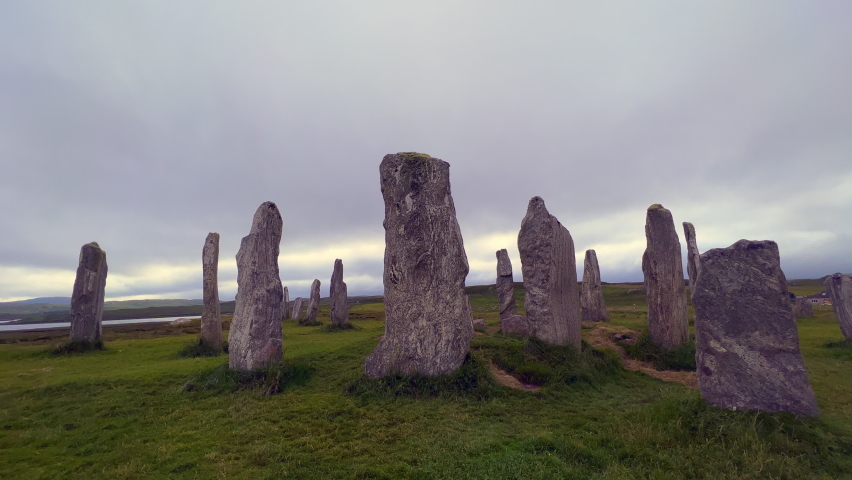 Moving around the standing stones or megaliths of the Callanish or Calanais stone circle in a cloudy evening. Sun peeking through the could, magic, ancient, neolithic atmosphere. Archaeological sites. Royalty-Free Stock Footage #1095885363