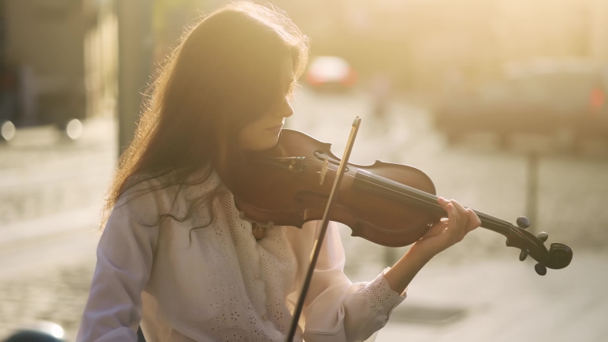 Young street musician playing classical wooden violin on the urban city street. Talented violinist with incredible sunset on the background.