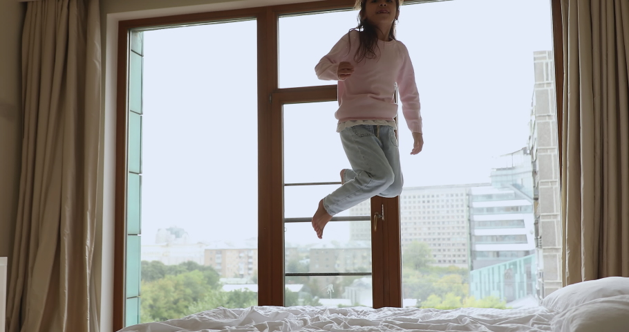 Energetic pretty girl kid jumping on comfortable spacious bed, with panoramic window in background, falling on soft mattress, white linen. Active child having fun at home, enjoying moving Royalty-Free Stock Footage #1095887207