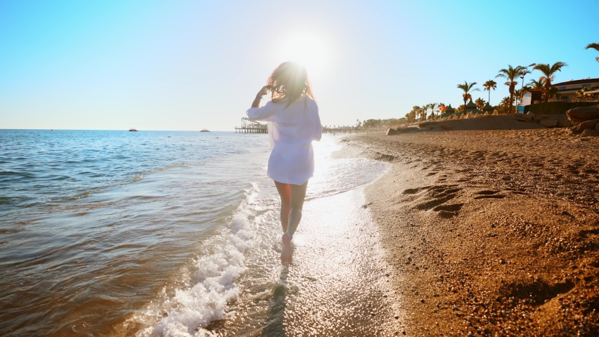 View from back of young woman on empty beach, who running on shore by sea and raises her arms up to sides in sense of freedom. Summer vacation and outdoors recreation. Happiness and relaxation.