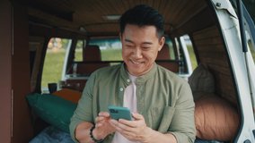 Footage of handsome joyful young Asian man holding smartphone and chatting. Smiling guy in olive shirt using gadget and sitting in cozy camping car. Weekend out of town. Outdoor