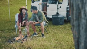 Shooting from behind tree. Cute happy couple in love talking, laughing and roasting marshmallows on skewers over fire pit. Date in nature. Candy-bouquet period in relationship. Leisure. Camping car - Powered by Shutterstock - Get 15% off with code: PIKWIZARD15