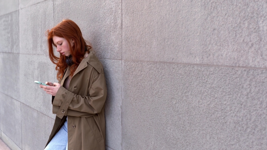 Cool teen hipster rebel girl with red hair standing at big city urban street wall background holding cell phone, using smartphone social media apps, shopping online in mobile phone application. Royalty-Free Stock Footage #1095898221