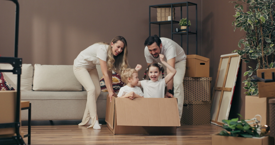 Happy family concept delighted daughter and son sitting in carton box playing with mother and father in new apartment moving in process concept. Royalty-Free Stock Footage #1095900455