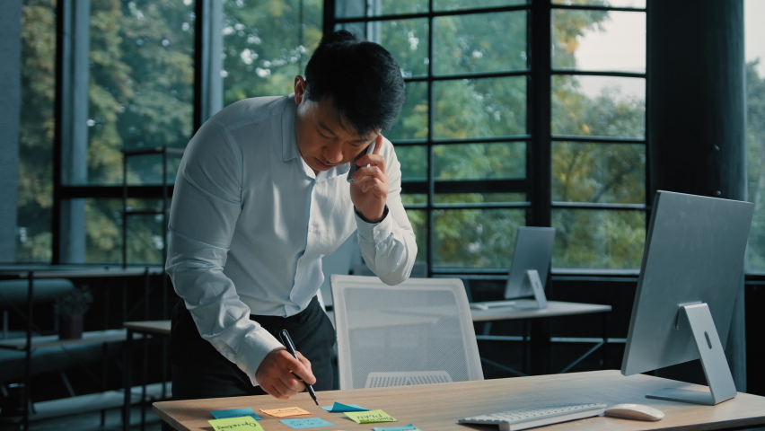 Busy asian businessman korean man working stand at table in modern office talk on phone give professional consultation by mobile call write tasks on sticky notes project paperwork multitasking concept | Shutterstock HD Video #1095902243