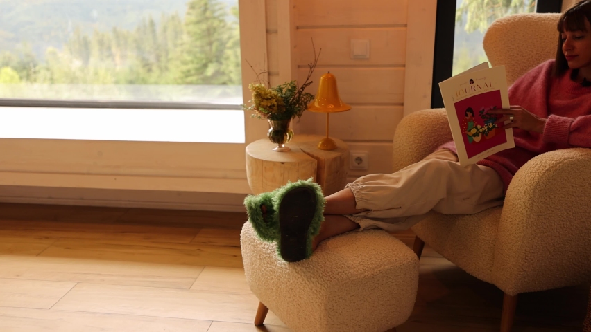 Woman reads some magazine while sitting in comfortable sofa in wooden house on nature. Home coziness, autumn time and domestic lifestyle concept | Shutterstock HD Video #1095913535