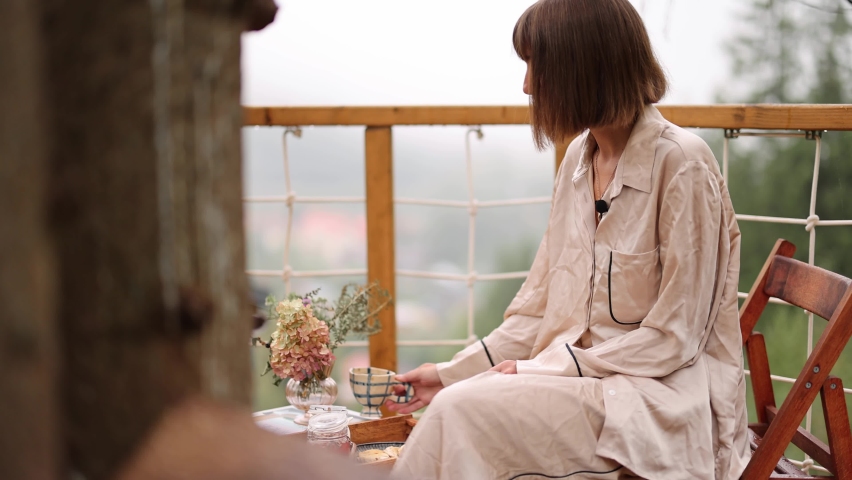 Woman in pajamas has a breakfast, drinking coffee on terrace of wooden house on tree in mountains. Cozy vacations on nature | Shutterstock HD Video #1095913541