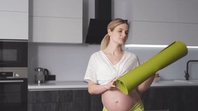 Happy cute pregnant woman standing with a fitness mat in her hands in her living room. Fitness for pregnant women.