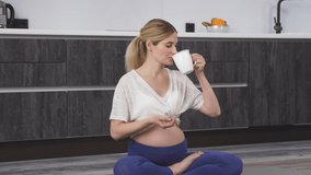 Pregnant happy woman enjoying a cup of water or tea, sitting on the floor after yoga at home, a woman in a tracksuit in her living room.