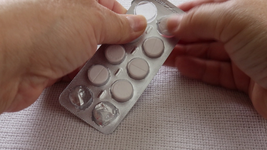 Female hands of an older adult woman taking two paracetamol tablets to relieve a headache caused by flu, white background.  Concept of large expenses for health services