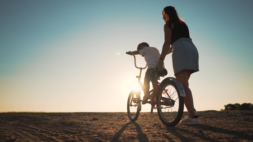 Happy family concept. Mom teaches her son to ride bike on green grass. Child rides bicycle along rural road. Green energy. Mom teaches her son to ride bike for the first time in park at sunset. Royalty-Free Stock Footage #1095915685