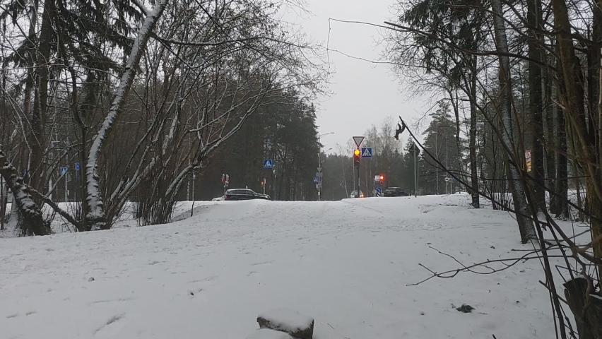The path extends to the edge of the city park and connects to the intersection of streets. There are traffic lights here, cars and pedestrians drive along the intersection. Ground is covered with snow