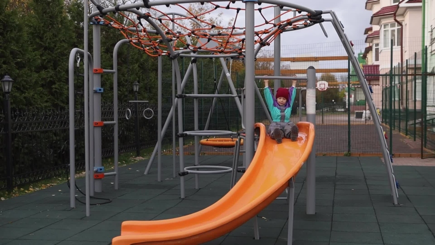 A boy in an autumn jumpsuit, hat and boots rides from a plastic slide.A child is playing on a walk in the playground.Active rest of the baby outside the house.The concept of a weekend with children