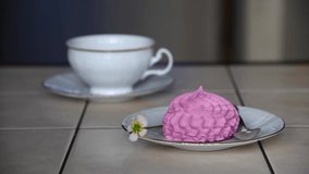 Pink meringue in white saucer lies on table with spoon and one flower. Blurred person pouring tea from teapot in white cup in the background. Selective focus. Real time video. Tea time theme.