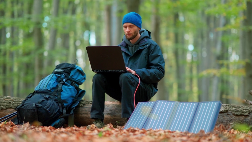 A young man goes hiking in the autumn forest. A solar panel charges a laptop in the forest.