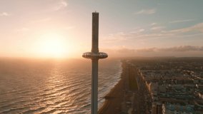Magical sunset 4k aerial video of British Airways i360 viewing tower pod with tourists in Brighton, UK with sea and Brighton Palace Pier in the background.