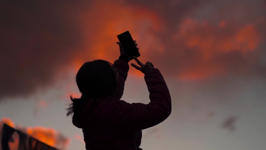 Silhouette of Indian female holding mobile phone and posing for selfie in background of sunset sky. Young Indian Travel Woman Vlogger vlogging during sunset in mountains.  