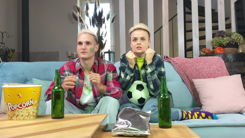 Young adult soccer fan couple with painted Brazilian flag on face cheering sport team watching live football match indoors. Stylish woman and happy man rejoicing celebrating goal sitting sofa at home