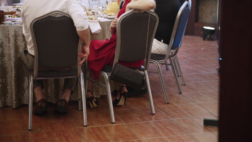 Unrecognizable woman in elegant red dress is trying to hang evening clutch on back of armchair at banquet table in restaurant. Chain falls down the back of chair, handbag lies on the floor.