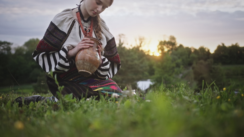 Woman in ethnic clothes pours milk from jug into jar on lawn Royalty-Free Stock Footage #1095939109