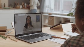 Over shoulder of laptop with video call of young female Caucasian online tutor in glasses smiling, showing papers, talking, waving hand to unrecognizable kid sitting by desk at home