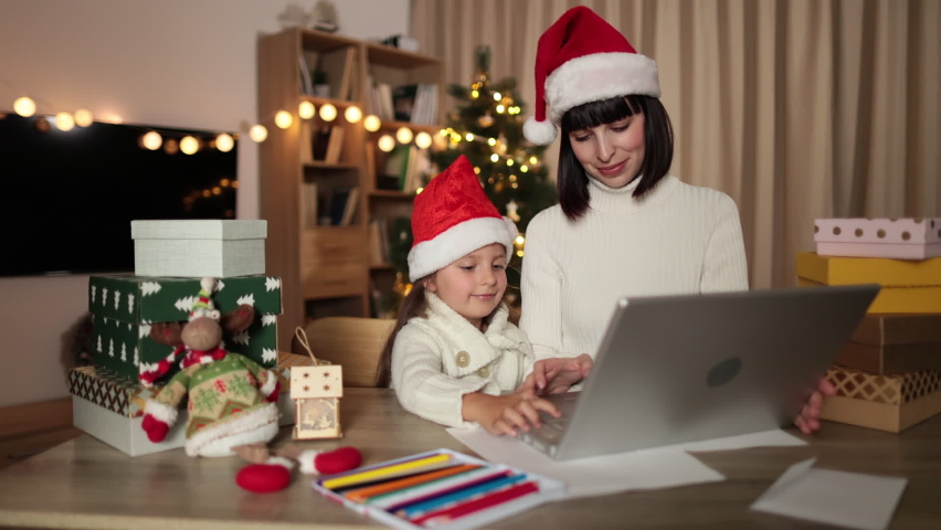 Indoor shot of beautiful happy young woman and her girl shopping online on laptop in cozy Christmas interior. Mother and daughter sitting at table with shopping gifts next to Christmas tree.