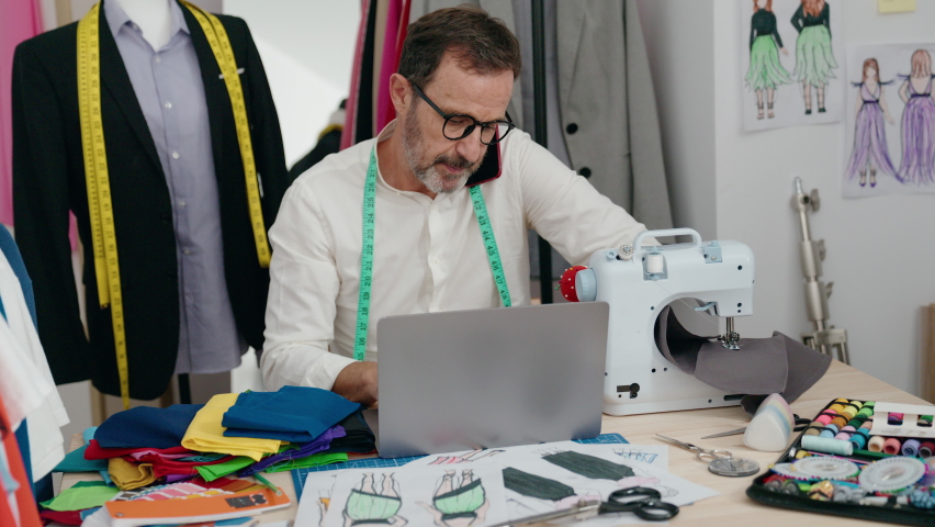 Middle age man tailor talking on smartphone using laptop at tailor shop
