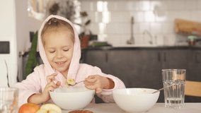 A little girl is eating porridge. A cute child dressed in a robe with a hood is sitting at a table and eating oatmeal porridge. close-up