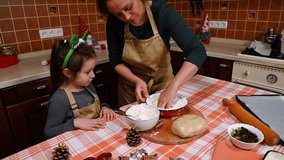 Beautiful multi-ethnic woman in green shirt and beige chef's apron, baker confectioner, pleasant housewife, loving mother, kneading cake dough with her adorable little daughter in the kitchen at home