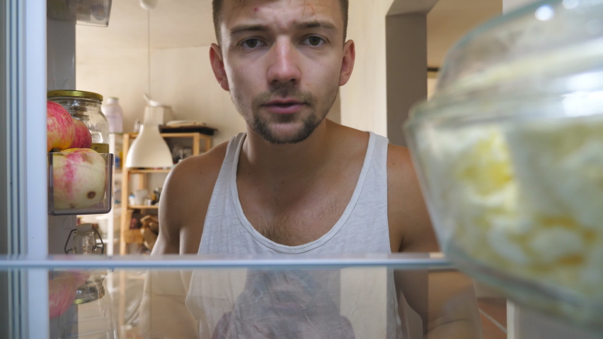 Young guy looking for food at refrigerator to has a breakfast at home. Sleepy man taking oiler and closing fridge at cuisine. Lazy human going to makes brunch at morning. Point of view Slow motion