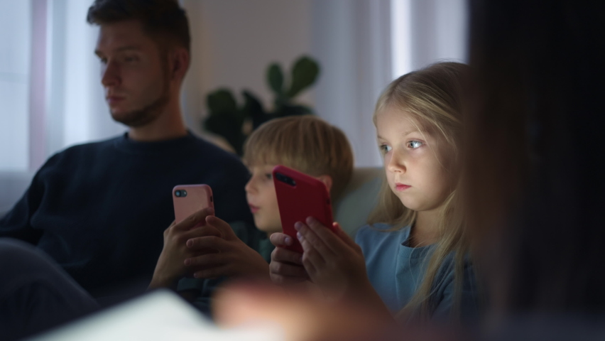 In the evening, two children sitting on the couch with their parents play mobile phones and do not communicate with the family. Phone addiction to games. Children and gadgets. Royalty-Free Stock Footage #1095970217