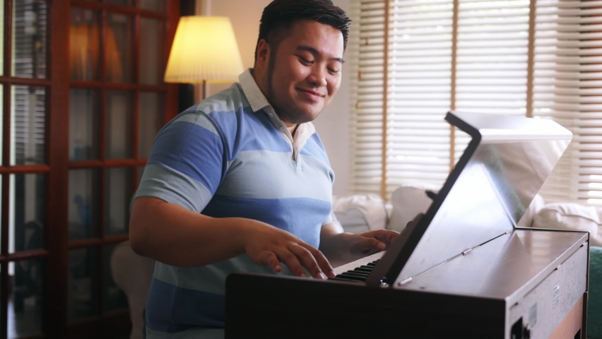 Young Asian chubby man singing and playing piano. Plus-size man practicing electric keyboard instrument at home. High quality 4k footage