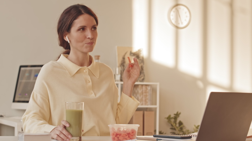 Young beautiful Caucasian woman eating healthy snacks and talking in wireless earphones while working from home